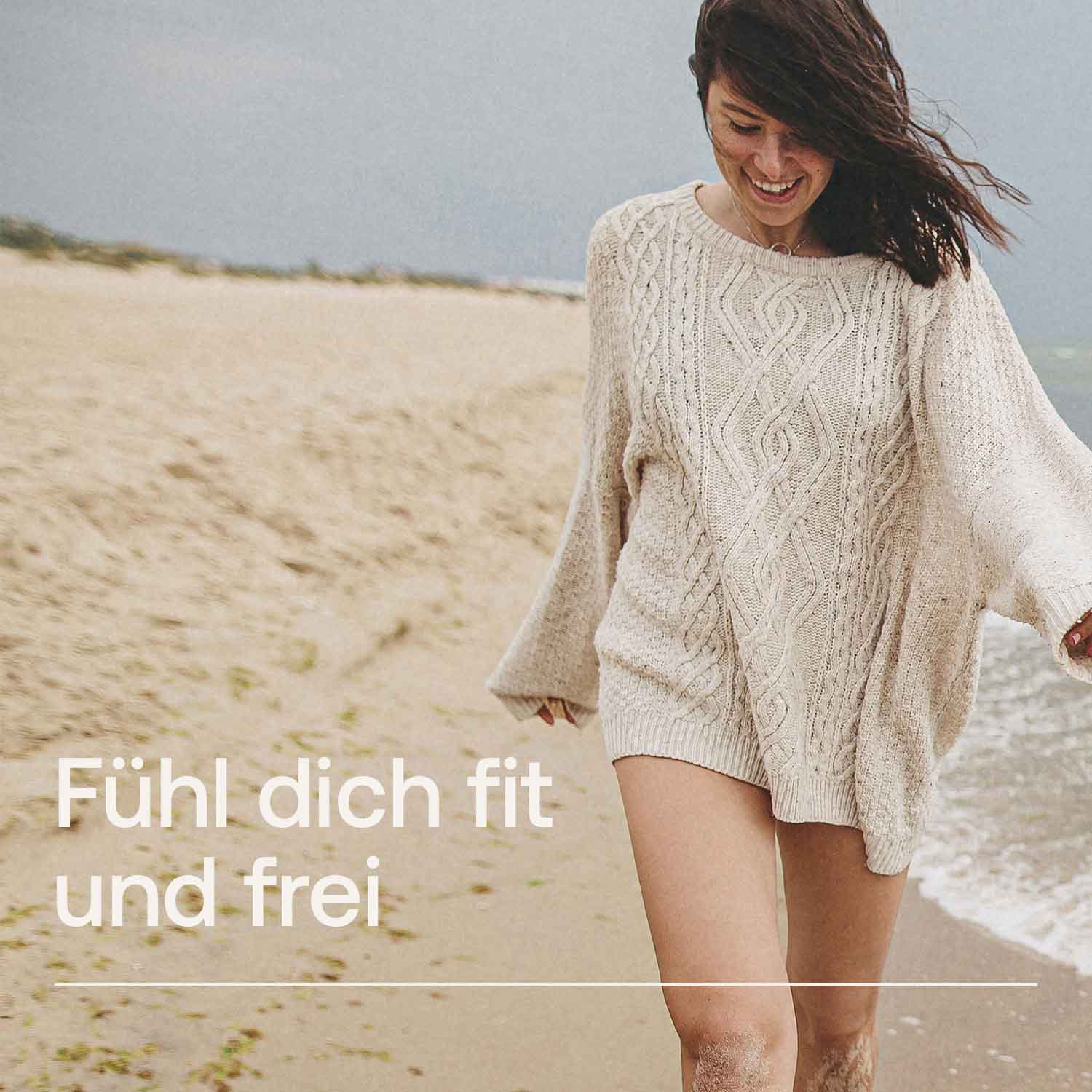 Lächelnde Frau im Strickpullover spaziert am Strand entlang mit Schriftzug 'Fühl dich fit und frei'