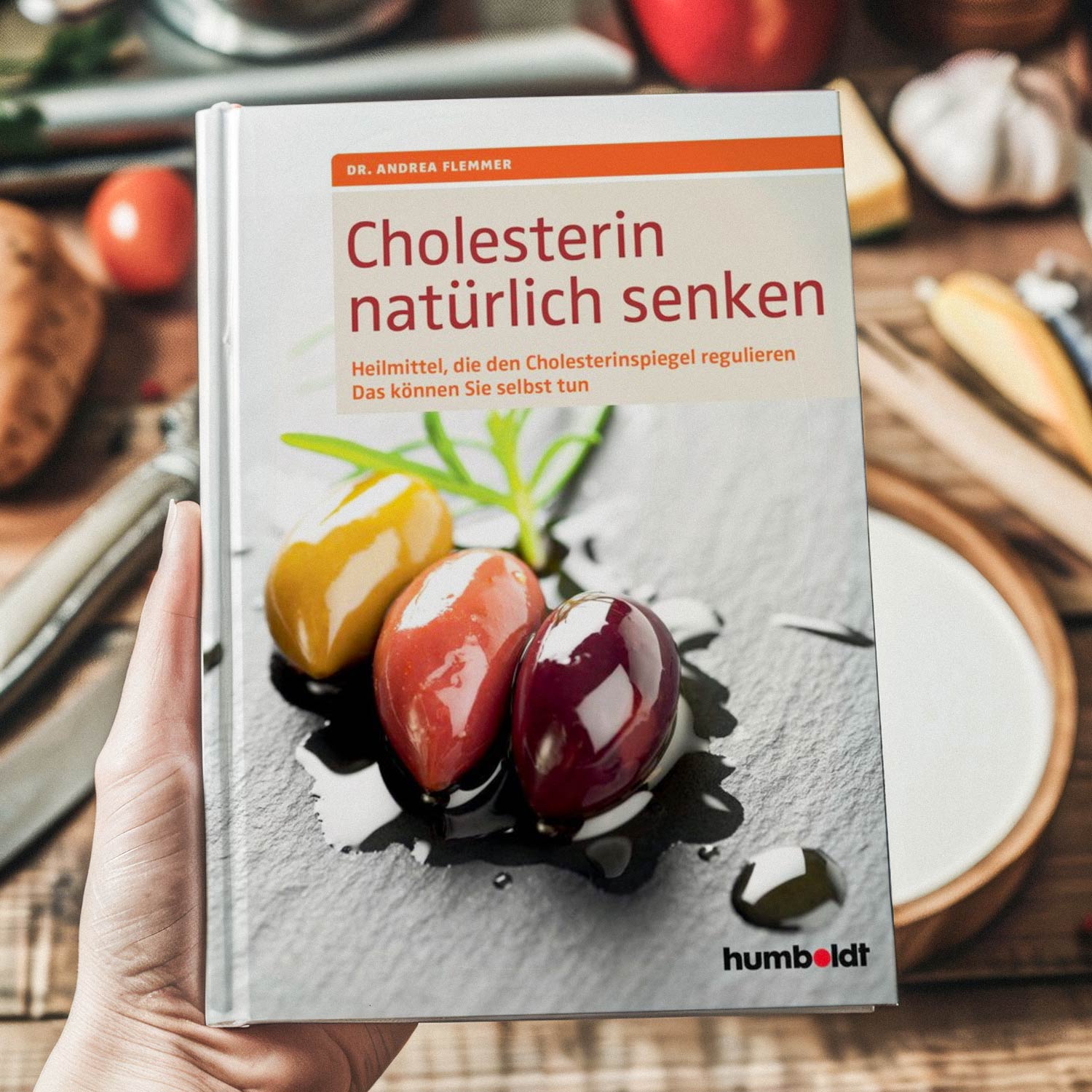 Hand hält das Buch 'Cholesterin natürlich senken' von Dr. Andrea Flemmer mit Oliven und Kochutensilien im Hintergrund