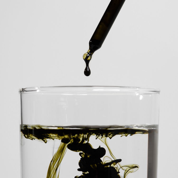 Nahaufnahme einer Pipette, die schwarze Flüssigkeit in ein Glas Wasser tropft, wodurch ein lebhaftes Muster entsteht.