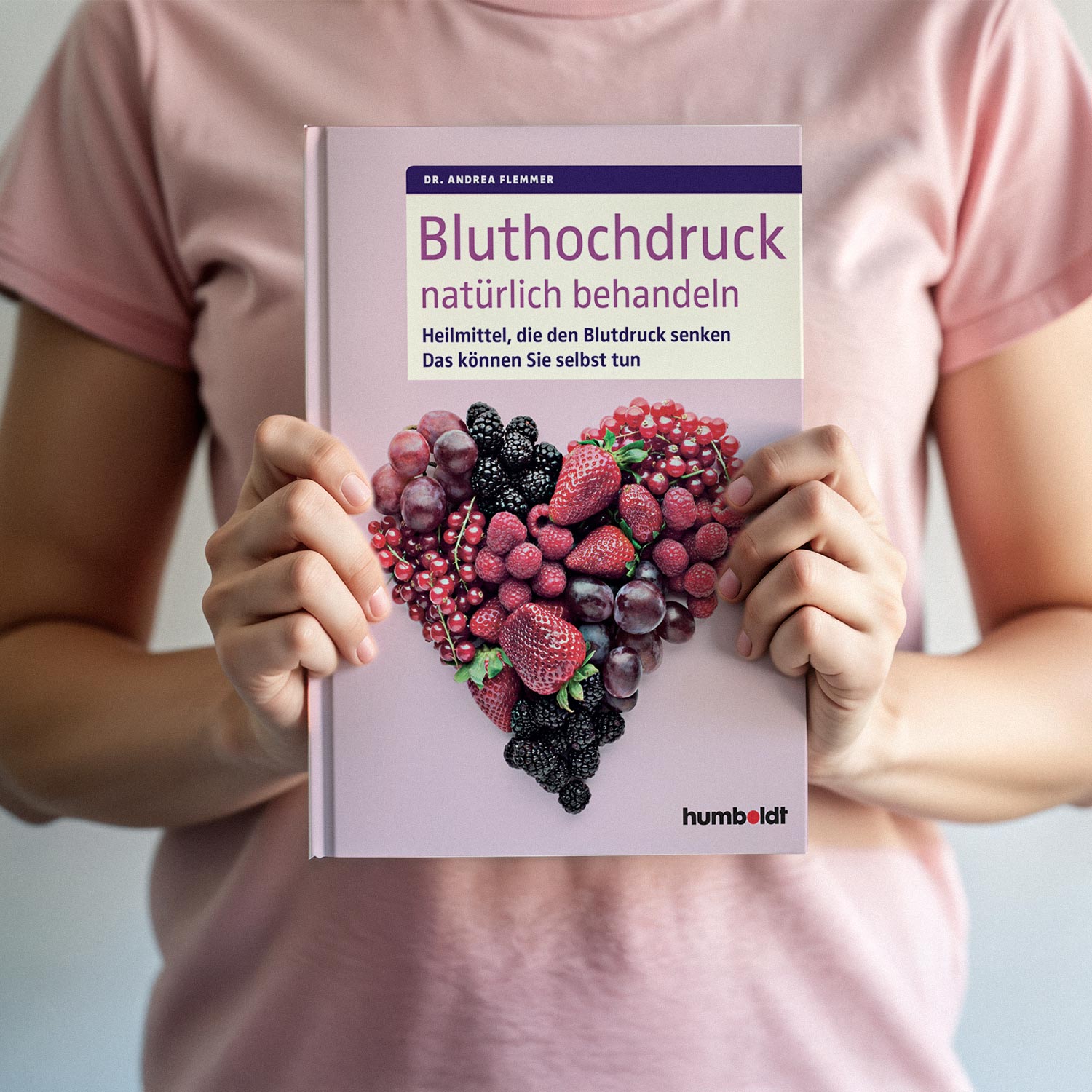 Person hält Buch mit dem Titel 'Bluthochdruck natürlich behandeln', illustriert mit einem Herz aus Beeren auf dem Cover